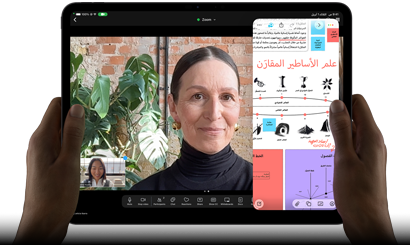 جهاز iPad Pro، الجزء الأمامي الخارجي، الوضع الأفقي، لون أسود فلكي، زوايا مدورة، إطار شاشة أسود، يدان تحملان الجهاز من الجانب الأيسر والأيمن، شاشة تعرض محادثة عبر فيس تايم تتم ترجمتها مباشرة، وتطبيق تعليمي يُظهر عرضاً تقديمياً بعنوان 'علم الأساطير المقارَن' مع نصوص ومخططات وملاحظات مكتوبة بخط اليد